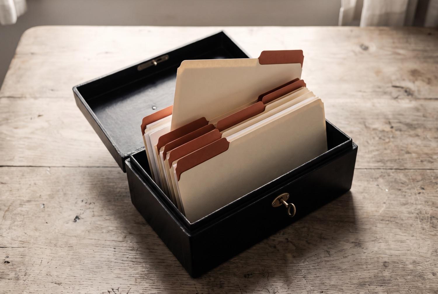 A black japanned deed box on a scrubbed table with its lid propped open, upright cream folders inside edged with terracotta tabs, one standing a little proud of the rest, and a small brass key in the lock.