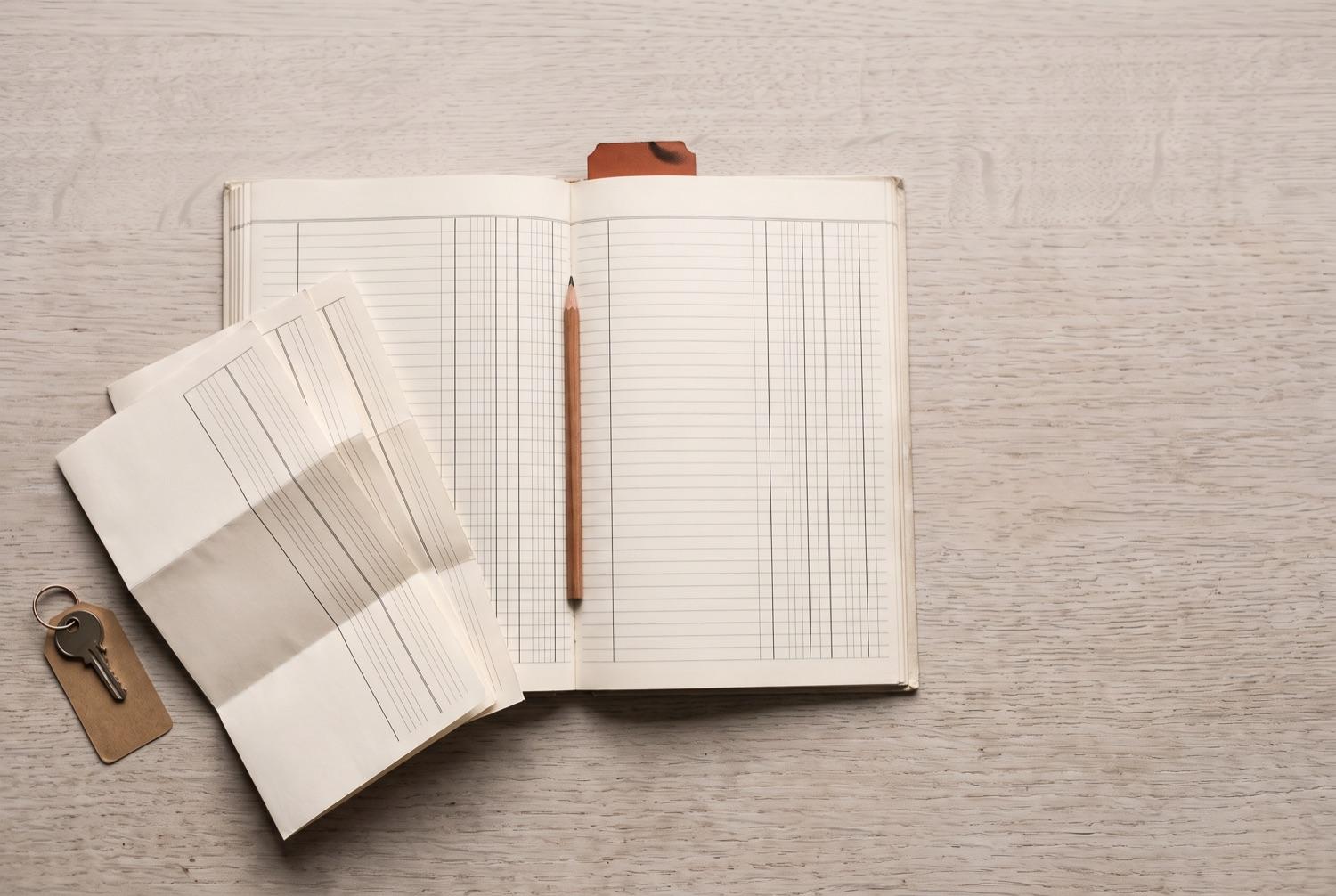 An open ledger showing blank ruled columns with a pencil resting in the gutter, statements folded in thirds and fanned beside it, a door key on a brown paper tag below them and a terracotta tab at the head of the page.