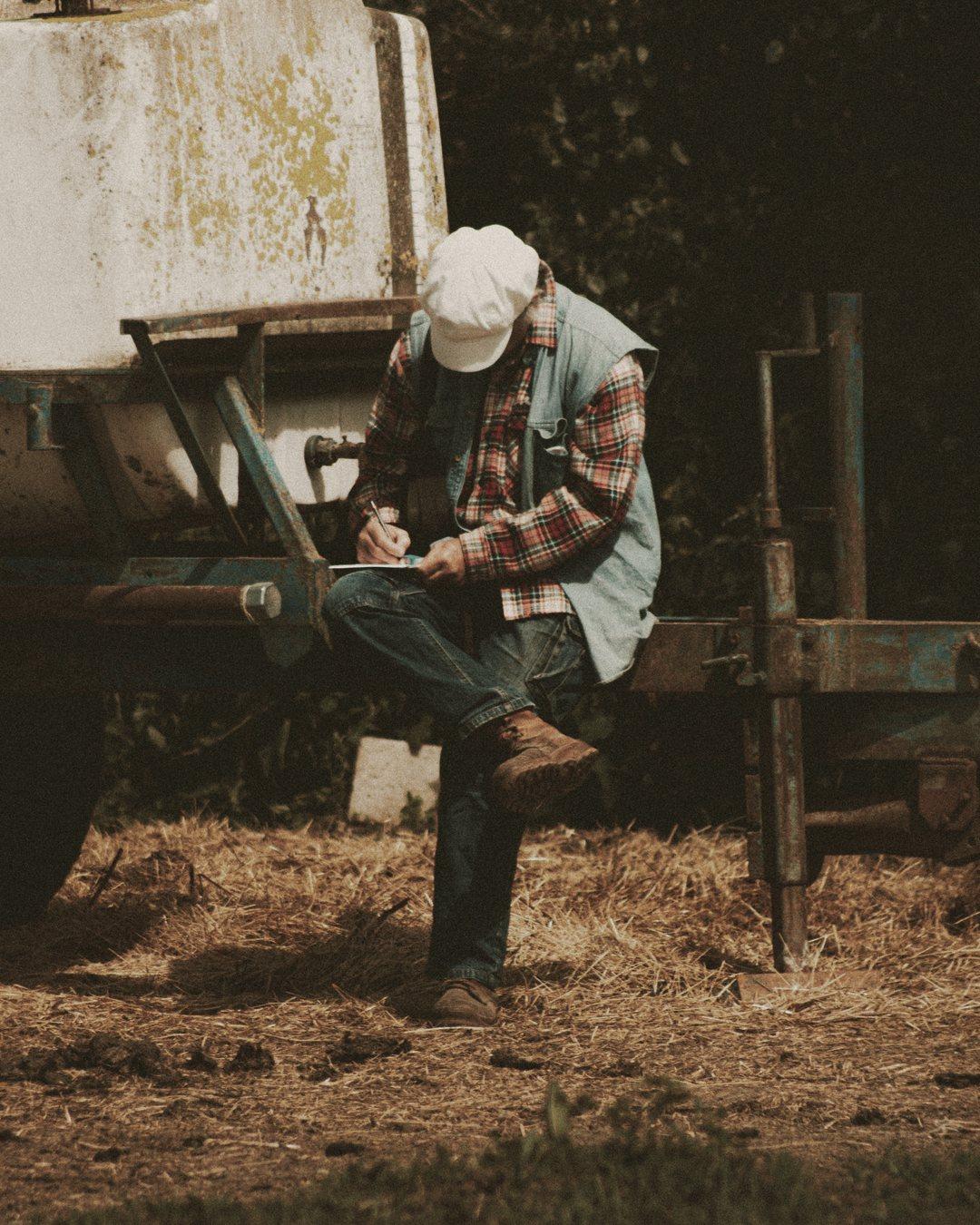 Farmer in a cap sitting on an old trailer, writing in a notebook