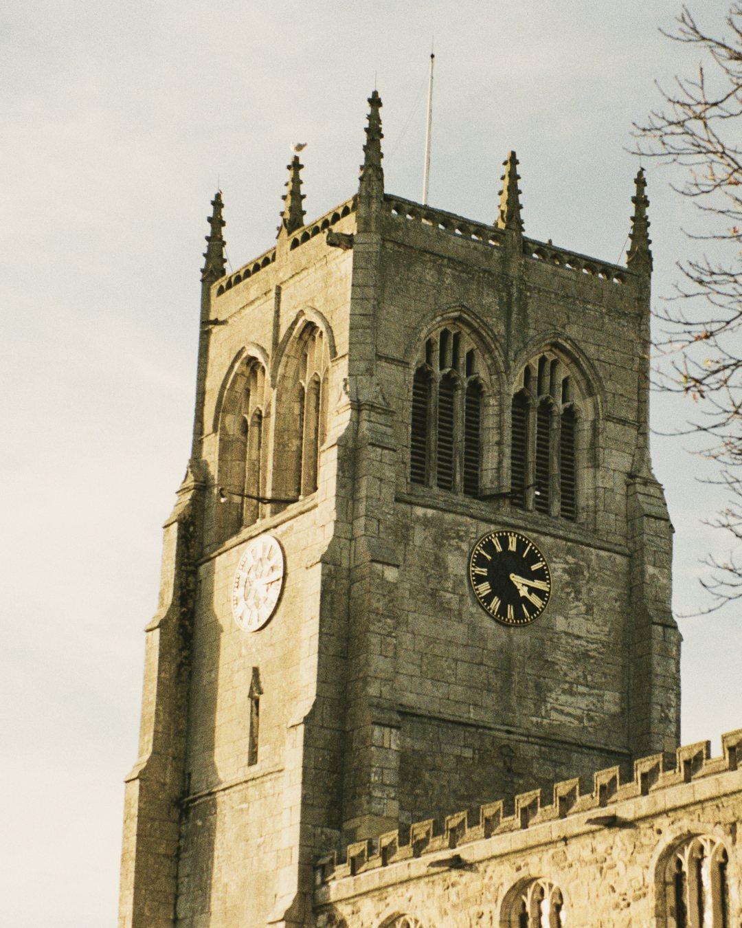 The tall stone tower of All Saints' Church in Preston, with two clock faces, beside an autumn tree
