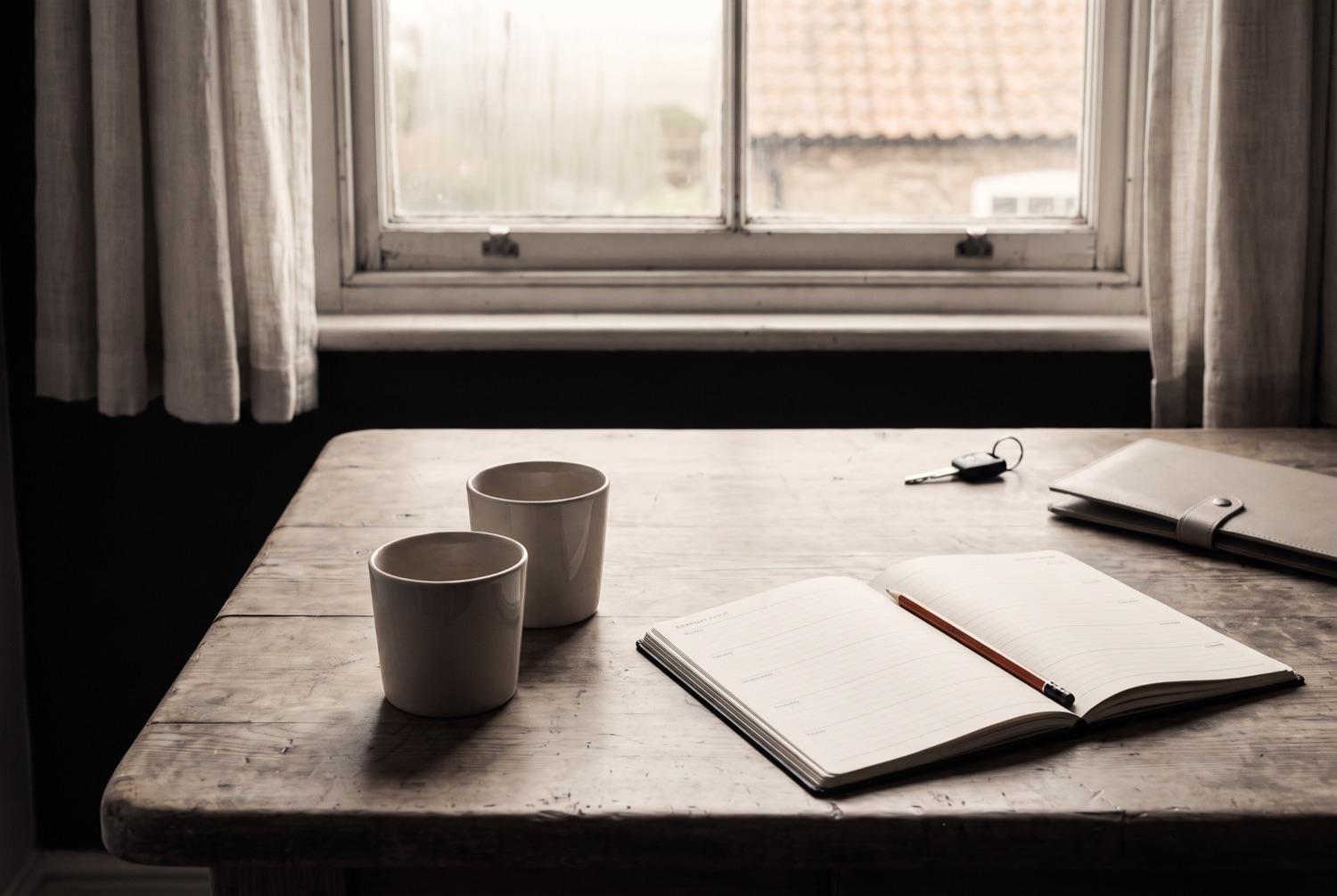 A kitchen table at a window with two cups the right way up, a diary open at a week of blank ruled lines with a terracotta pencil in the gutter, and a car key and a closed document wallet at the far edge.