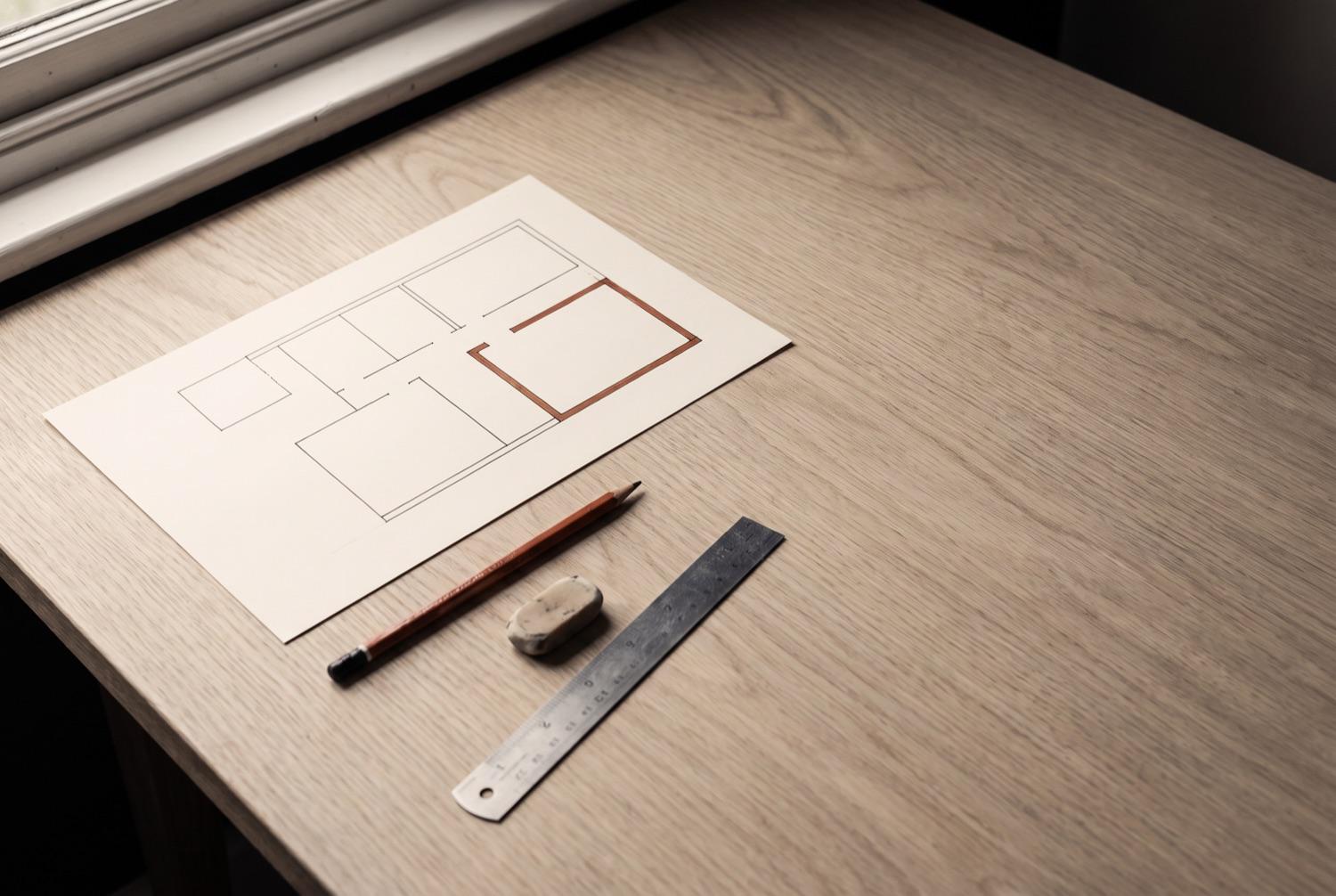 A sheet of cream paper on a bare desk carrying a hand drawn diagram of empty boxes joined by ruled ink lines, one box outlined in terracotta, with a pencil, a soft eraser and a steel rule lying below it.
