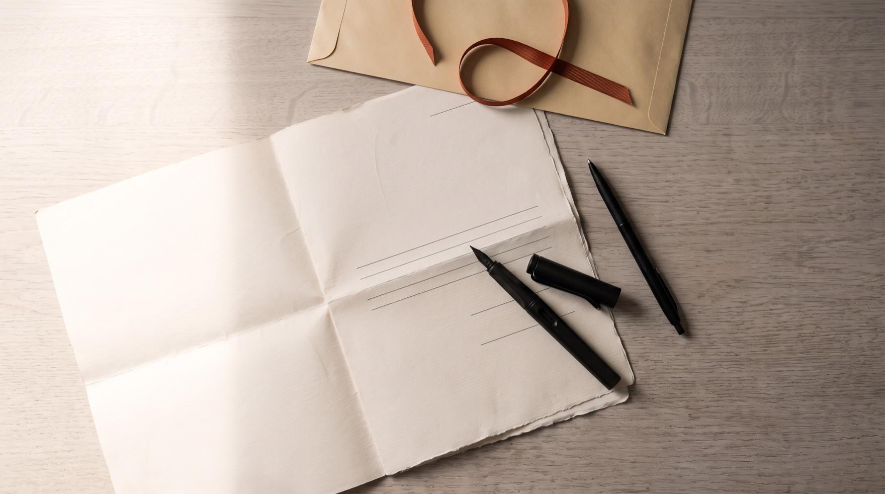 Seen from above, a creased document lying open at a panel of blank ruled lines, a black fountain pen with its cap off and a plain ballpoint set down across it, and a long unaddressed manila envelope above them with a terracotta ribbon curled loose on it.